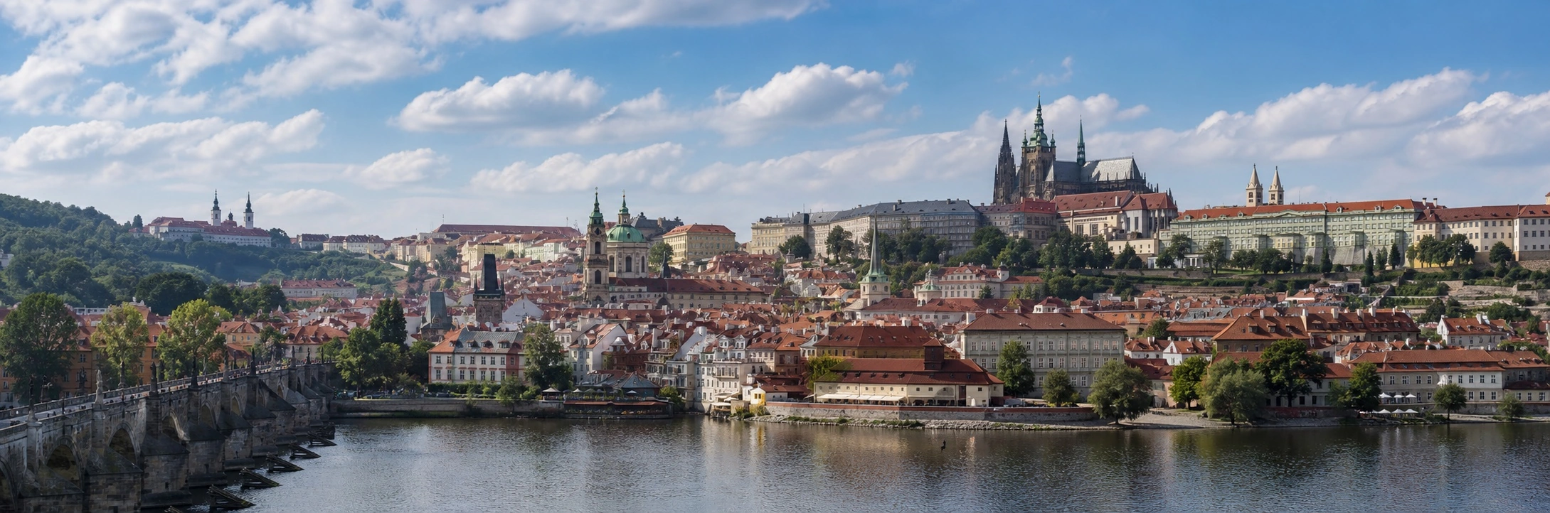 Prague Castle and St. Vitus Cathedral above the Vltava River and Charles Bridge beneath a blue sky.